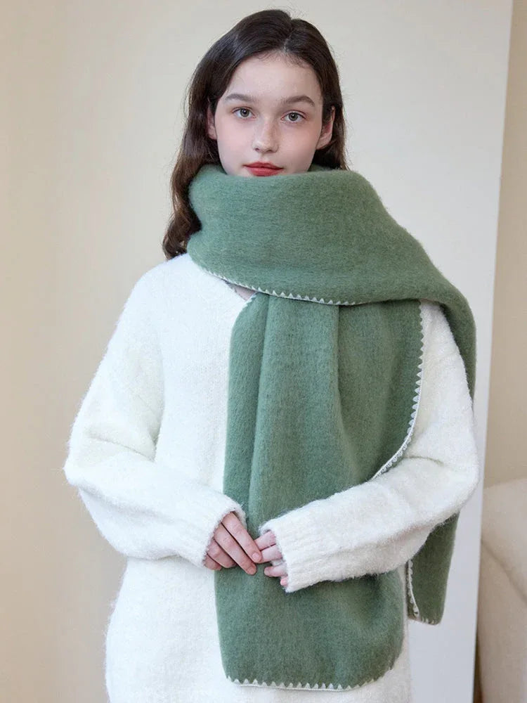 Woman wearing an oversized green scarf with a white fringed edge and a cream-colored sweater, with hands clasped in front.