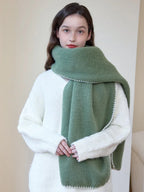 Woman wearing an oversized green scarf with a white fringed edge and a cream-colored sweater, with hands clasped in front.