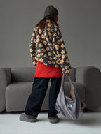 Woman wearing a grey fleece jacket with a yellow floral print, black pants, a red top, and carrying a grey tote bag.