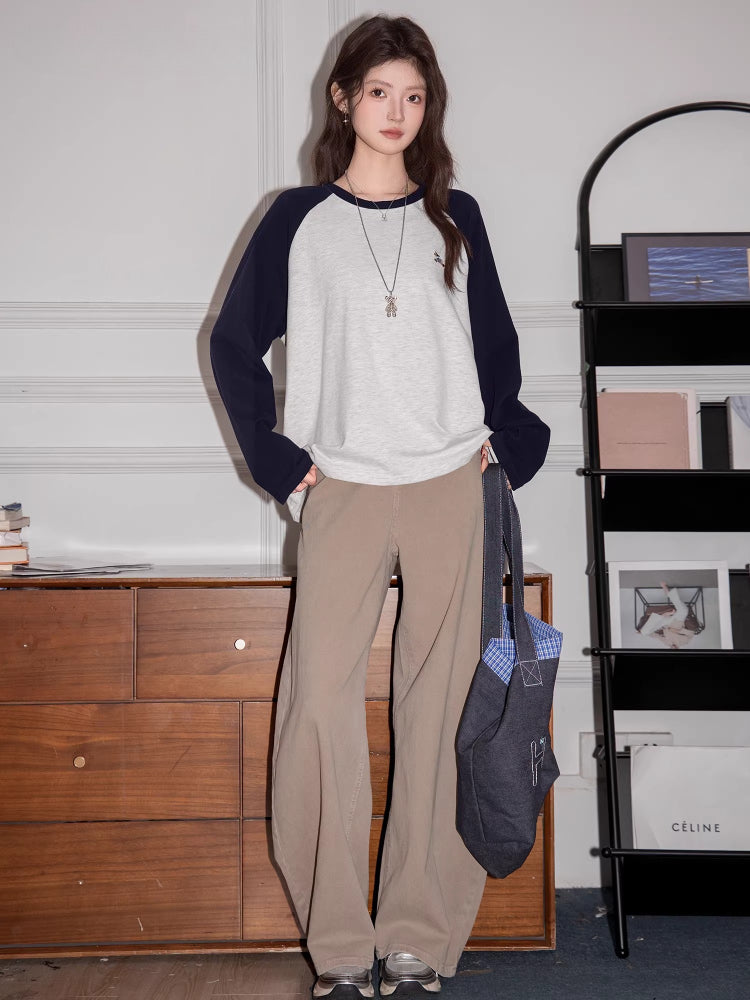 Full body shot of a woman wearing a light grey long-sleeved t-shirt with navy blue raglan sleeves and beige pants, holding a dark blue tote bag.