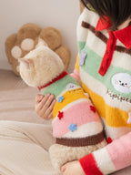 A person holds a cream-colored cat wearing a colorful striped sweater, with the cat's head turned to the side.