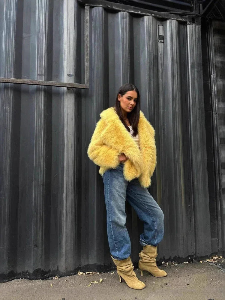 Full body shot of a woman in a yellow faux fur jacket, blue jeans, and beige boots, posing against a corrugated metal wall.
