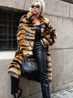 Woman wearing a long faux fur coat with a tiger stripe print, black pants, and a black handbag, leaning against a textured wall.