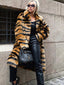 Woman wearing a long faux fur coat with a tiger stripe print, black pants, and a black handbag, leaning against a textured wall.