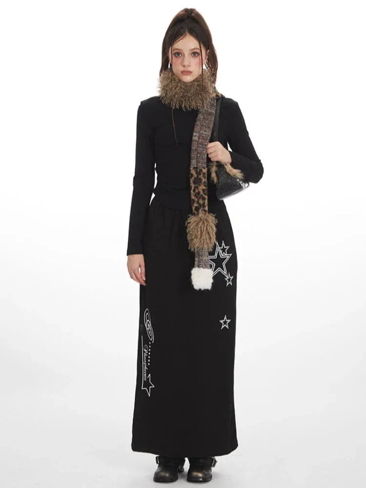 Model wearing a black long-sleeve top and a black maxi skirt with a drawstring waist and white star graphics, accessorized with a multi-patterned scarf and a black handbag.