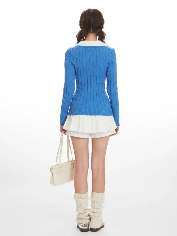 Woman wearing a blue cable knit V-neck sweater with a white collar over a white pleated skirt, cream leg warmers, and a cream handbag, back view.
