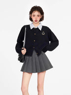 Woman wearing a black knit cardigan over a striped collared shirt, paired with a grey pleated skirt and a black backpack.