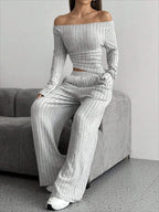Model sitting on a sofa wearing a grey ribbed knit off-the-shoulder long sleeve top and wide leg pants set.