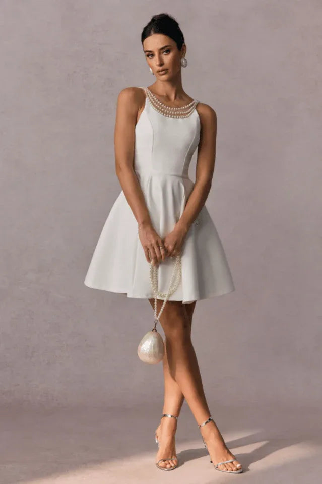 Full length shot of a woman in a short white A-line dress with pearl details, holding a pearl handbag
