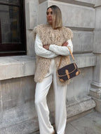 Woman wearing a beige shaggy faux fur vest over a white cable-knit sweater and white wide-leg pants, with arms crossed and a brown and tan crossbody bag.