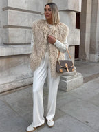 Woman wearing a beige shaggy faux fur vest over a white cable-knit sweater and white wide-leg pants, with a brown and tan crossbody bag and white sneakers.