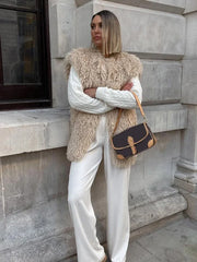Woman wearing a beige shaggy faux fur vest over a white cable-knit sweater and white wide-leg pants, with a brown and tan crossbody bag.