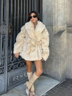 woman wearing a cream faux fur coat with a large collar and a belt, standing with legs apart