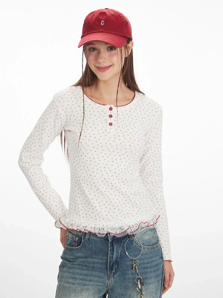Close-up of a model wearing a white long-sleeve top with a floral print and red trim, paired with blue jeans and a red baseball cap.
