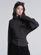 Woman in a dark grey turtleneck sweater with batwing sleeves and a cinched button detail at the waist, with sunglasses on her head.