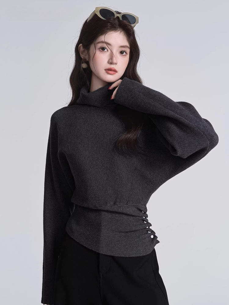 Woman in a dark grey turtleneck sweater with batwing sleeves and a cinched button detail at the waist, with sunglasses on her head.