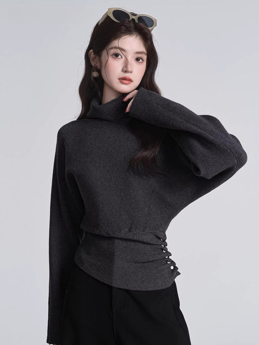 Woman in a dark grey turtleneck sweater with batwing sleeves and a cinched button detail at the waist, with sunglasses on her head.