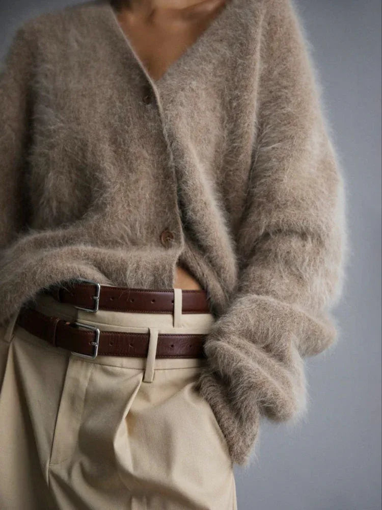 Model wearing a beige fuzzy V-neck button-up cardigan with tucked-in hands and light brown pants with a double belt detail.