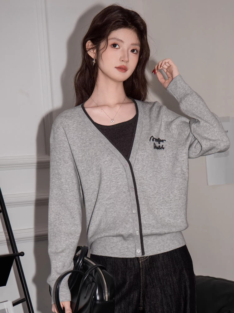 Woman holding a black handbag, wearing a grey knit cardigan with black trim and embroidery, dark top, and dark wide-leg jeans, sitting on a black couch.