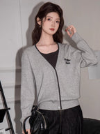 Woman holding a black handbag, wearing a grey knit cardigan with black trim and embroidery, dark top, and dark wide-leg jeans, sitting on a black couch.