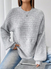 Close-up of a woman wearing a light grey oversized knit sweater with balloon sleeves and a crew neck, paired with blue denim jeans and a silver heart pendant necklace.