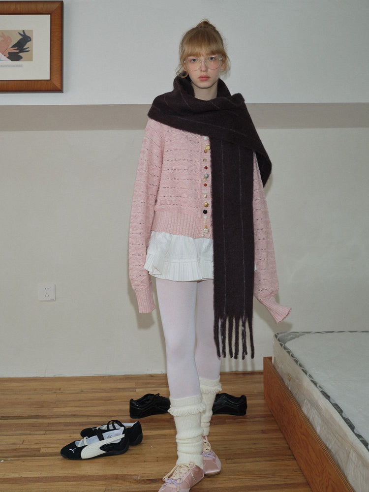A young woman stands with her arms at her sides, wearing a pink knit cardigan over a white pleated skirt, a long dark scarf, white tights, leg warmers, and pink ballet flats.