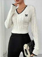 Woman wearing a white cable knit cropped V-neck sweater with black trim and a black bow detail, paired with black pants and a black handbag.