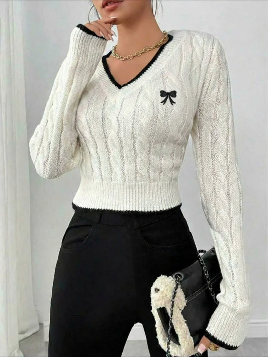 Woman wearing a white cable knit cropped V-neck sweater with black trim and a black bow detail, paired with black pants and a black handbag.