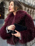 Woman wearing a burgundy shaggy faux fur cropped jacket and holding a black clutch bag.