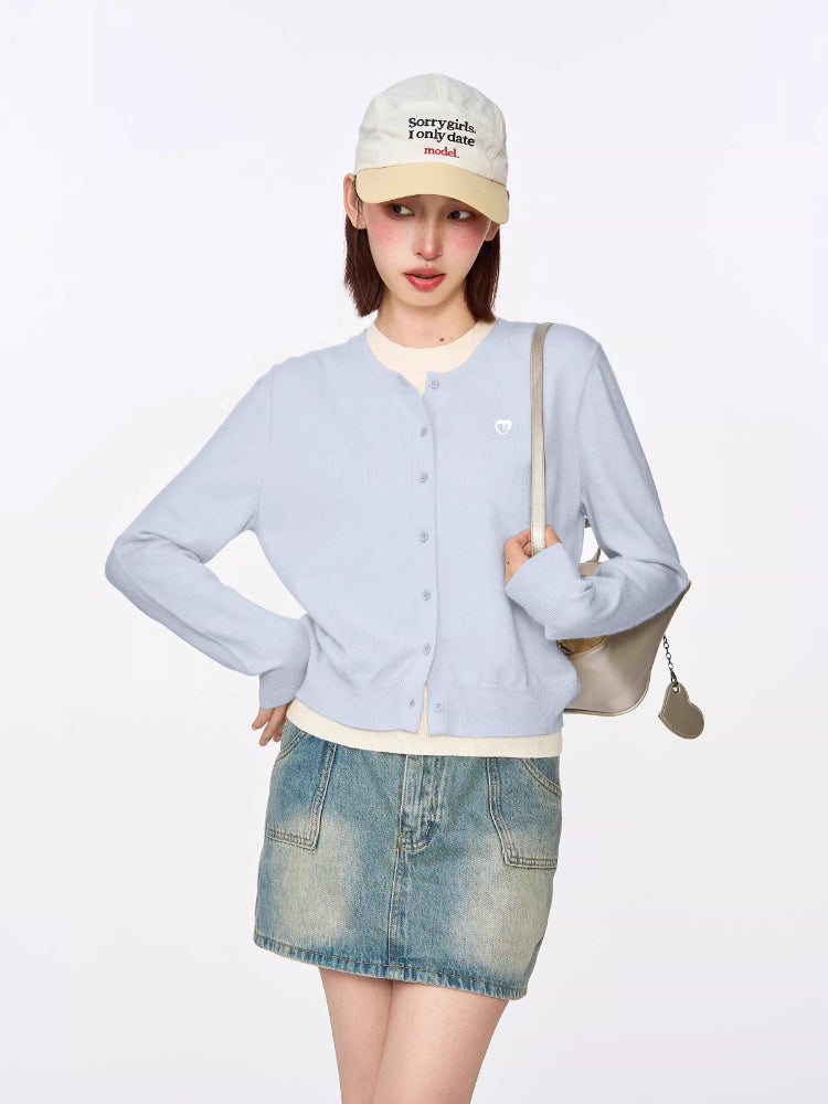 Front view of a model in a light blue cropped cardigan, white top, and denim mini skirt, holding a silver handbag.