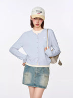 Front view of a model in a light blue cropped cardigan, white top, and denim mini skirt, holding a silver handbag.