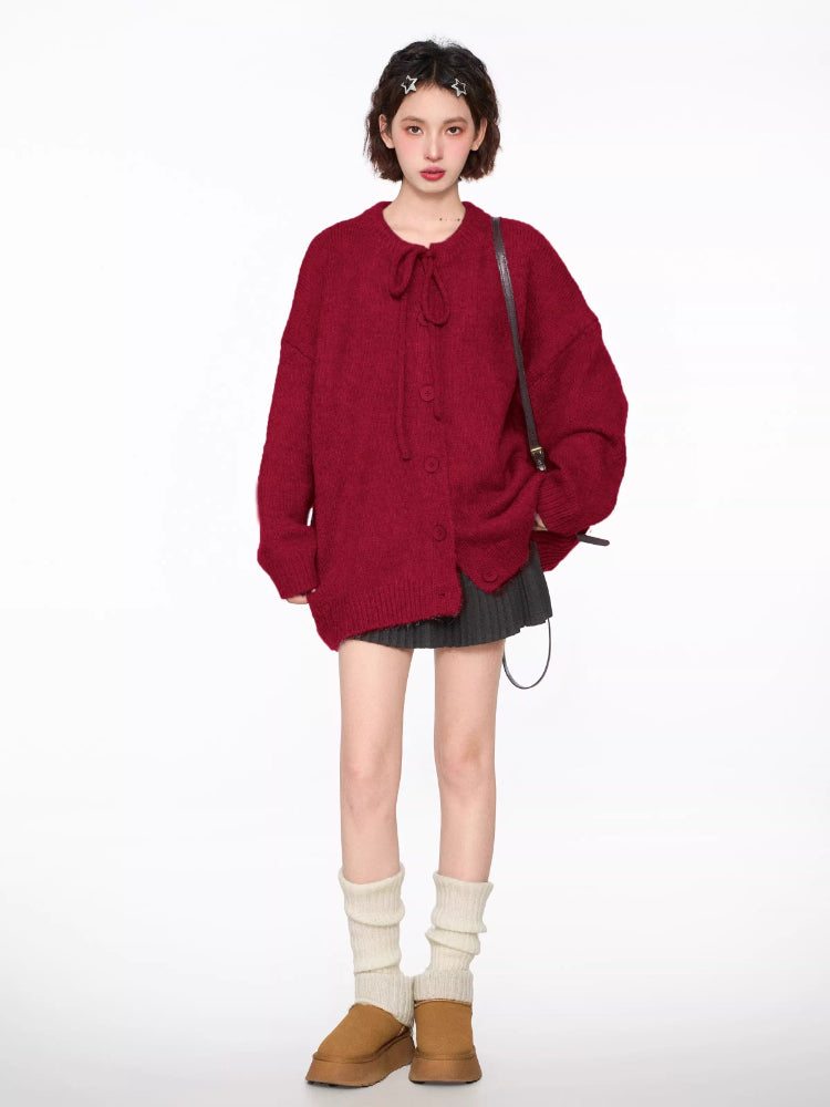 Full body shot of a woman wearing a red knit cardigan with tie neck and buttons, grey pleated skirt, brown crossbody bag, and white leg warmers.