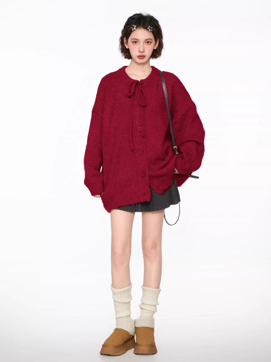 Full body shot of a woman wearing a red knit cardigan with tie neck and buttons, grey pleated skirt, brown crossbody bag, and white leg warmers.