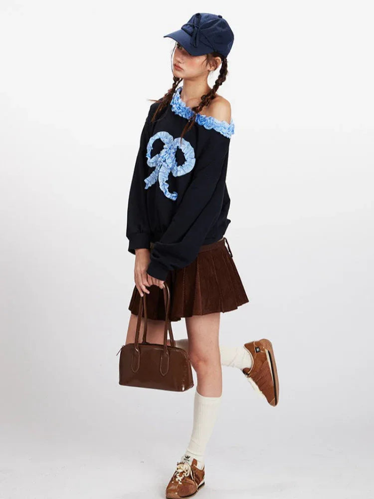 Full-body shot of a model posing in a black off-the-shoulder sweatshirt with a blue bow, brown pleated skirt, and white socks.