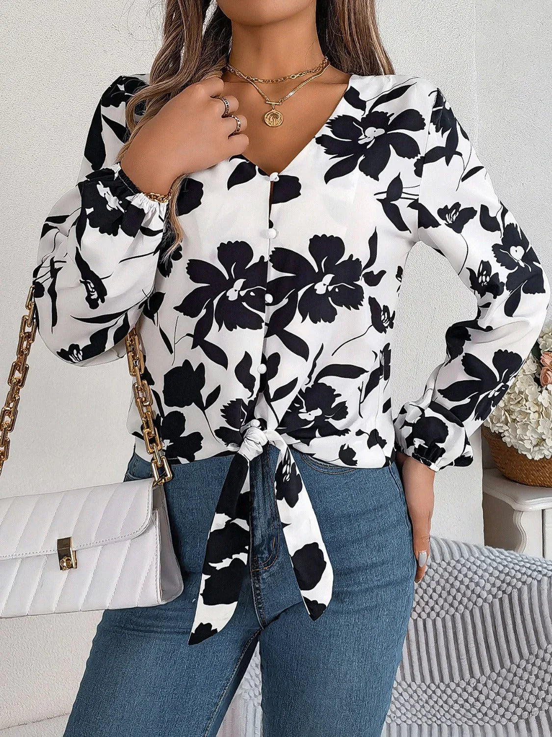 Woman wearing a black and white floral print long sleeve blouse with a front tie detail and blue jeans, holding a white handbag, front view