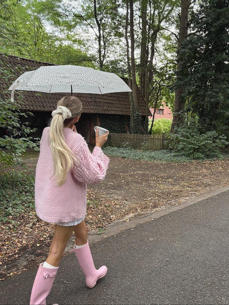 Rear view of a woman in a pink cable knit sweater, white shorts, and pink rain boots, walking with a poodle, holding a mug and an umbrella.