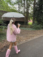 Rear view of a woman in a pink cable knit sweater, white shorts, and pink rain boots, walking with a poodle, holding a mug and an umbrella.
