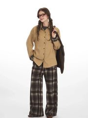 Full-body shot of a model in a tan button-down shirt and brown plaid wide-leg pants, carrying a brown knitted bag.