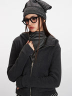 Close-up front view of a woman wearing a charcoal grey hooded zip-up jacket over a black and white striped turtleneck, with her hand on her chest.