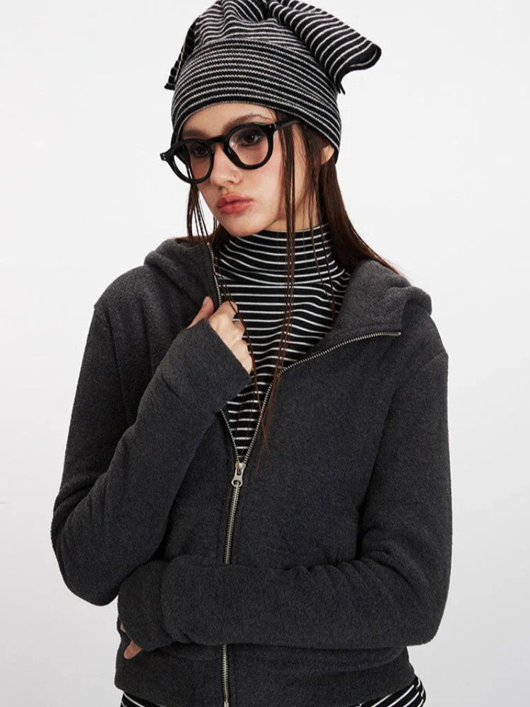 Close-up front view of a woman wearing a charcoal grey hooded zip-up jacket over a black and white striped turtleneck, with her hand on her chest.