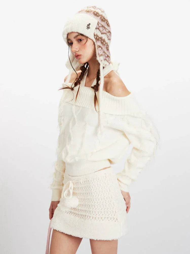 Side view close-up of a model wearing a cream off-the-shoulder knit sweater and a matching mini skirt, with an ear flap hat and pink handbag.