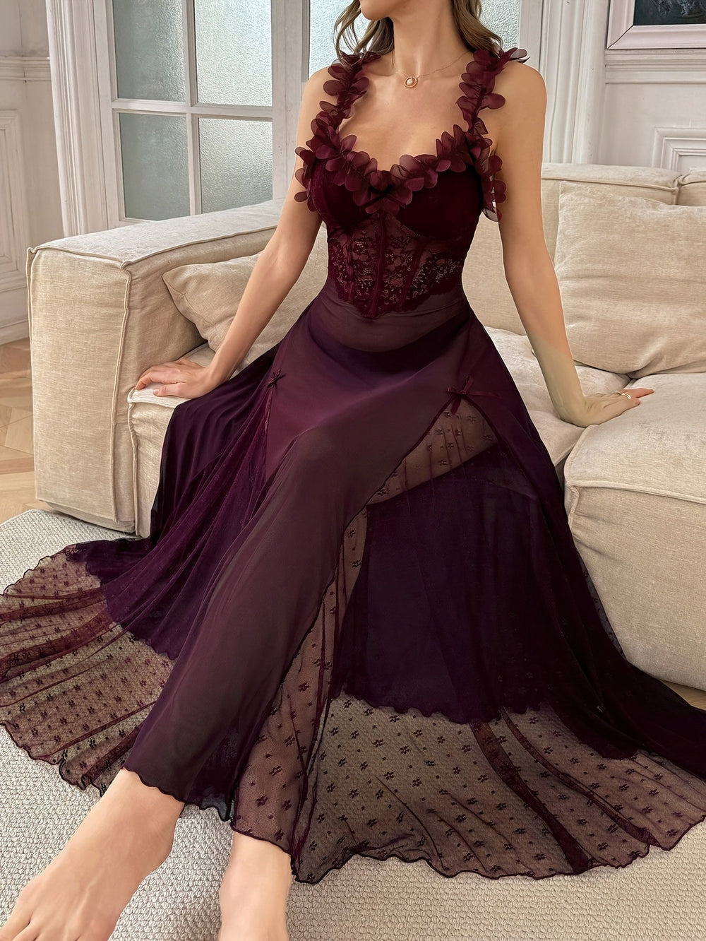 Woman wearing a sheer plum maxi slip dress with floral applique straps sitting on a cream sofa.