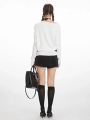 Back view of a model wearing a white off-the-shoulder long-sleeve top, black high-waisted shorts, black knee-high socks, and black heels, holding a black tote bag.