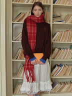 A young woman stands in front of a bookshelf wearing a brown sweatshirt, grey cycling shorts, and a blue and white plaid scarf.