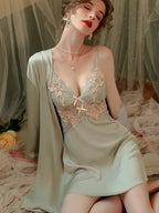 Woman in a light green satin slip dress with lace detailing and a matching robe, seated on a bed with floral accents.