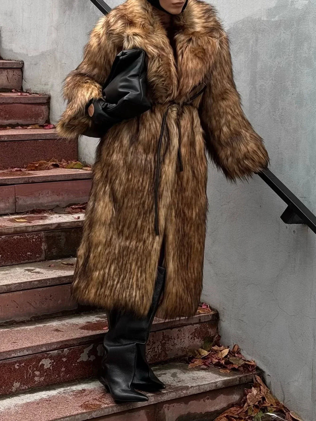 Woman wearing a long brown faux fur coat with a black scarf, black boots, and a black bag, standing on stairs.
