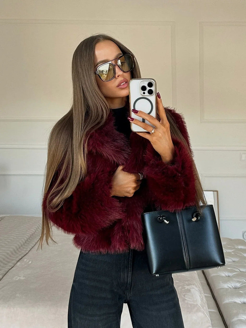 Woman holding a black handbag and adjusting her burgundy faux fur jacket while taking a selfie