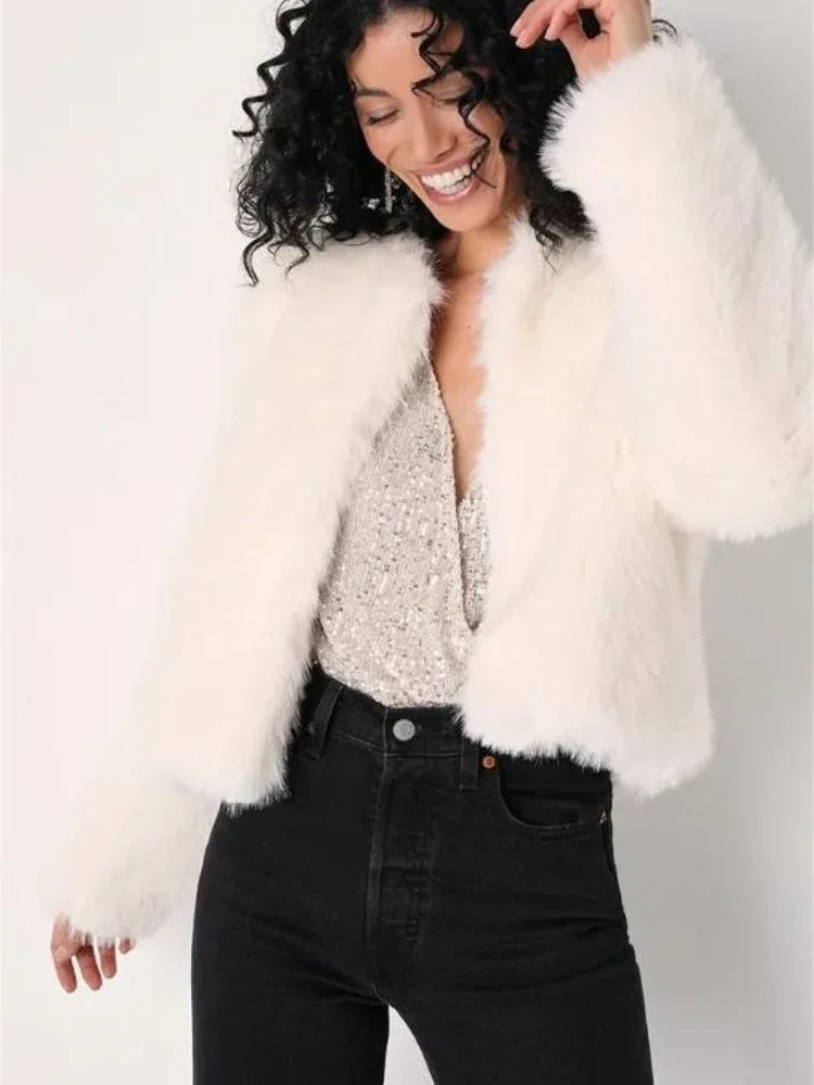Woman with curly hair smiling, wearing a white faux fur jacket over a sparkly top and black jeans.