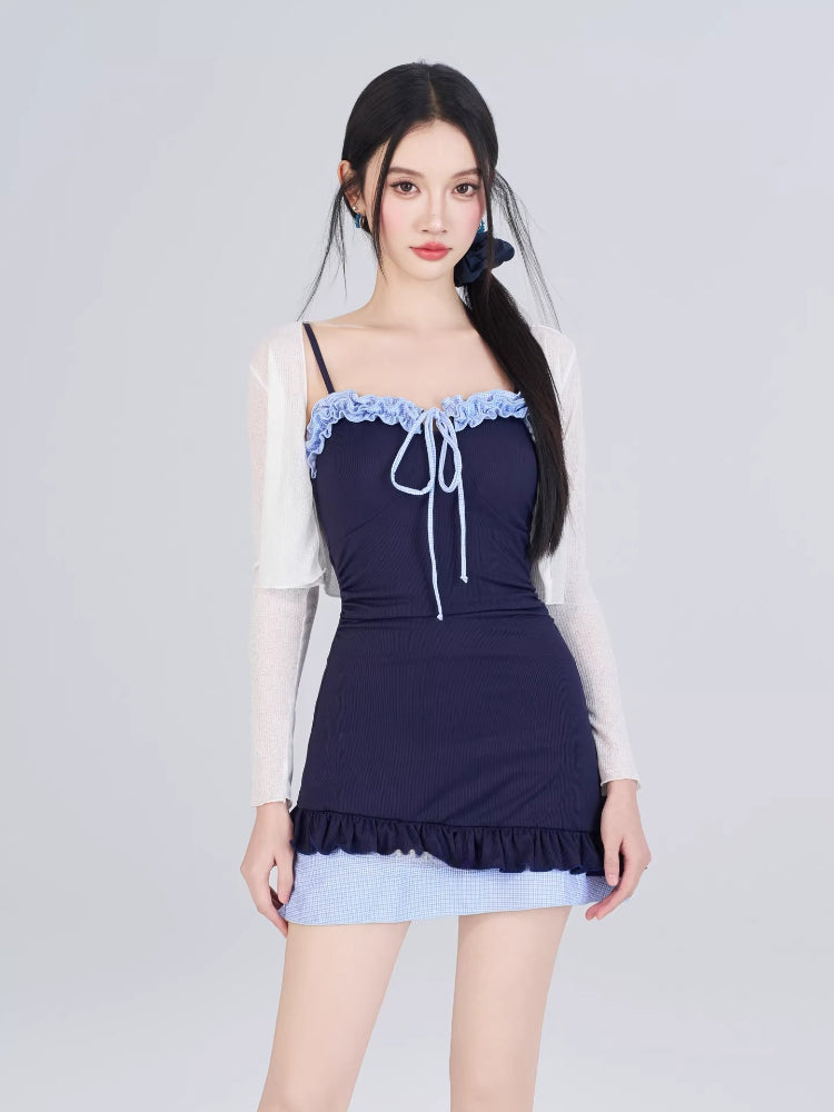 Model wearing a navy blue ribbed knit one-piece swimsuit with light blue gingham trim and a white sheer long-sleeved cardigan, with hair tied back.