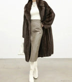 Woman wearing a brown mink fur coat, a white turtleneck, a grey leather skirt, and white boots, holding a white handbag.